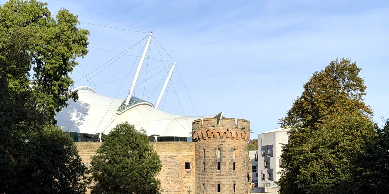 An old stone tower among trees, with the white tent roof of Dynamic Earth behind it